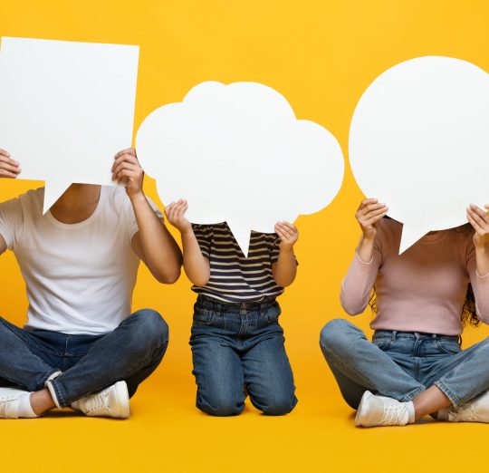 Diverse Thoughts. Father, Mother And Little Daughter Holding Different Empty Speech Bubbles In Front Of Their Faces, Unrecognizable Family Sitting On Floor On Yellow Studio Background, Copy Space